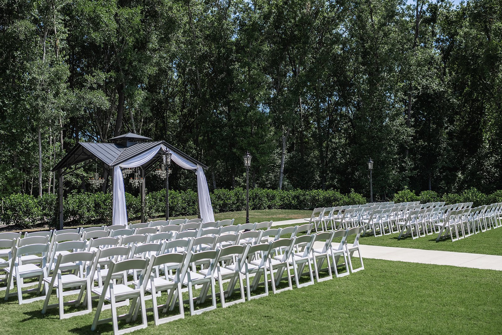 Outdoor wedding ceremony setup with white chairs arranged in rows facing a decorated gazebo under tall trees.