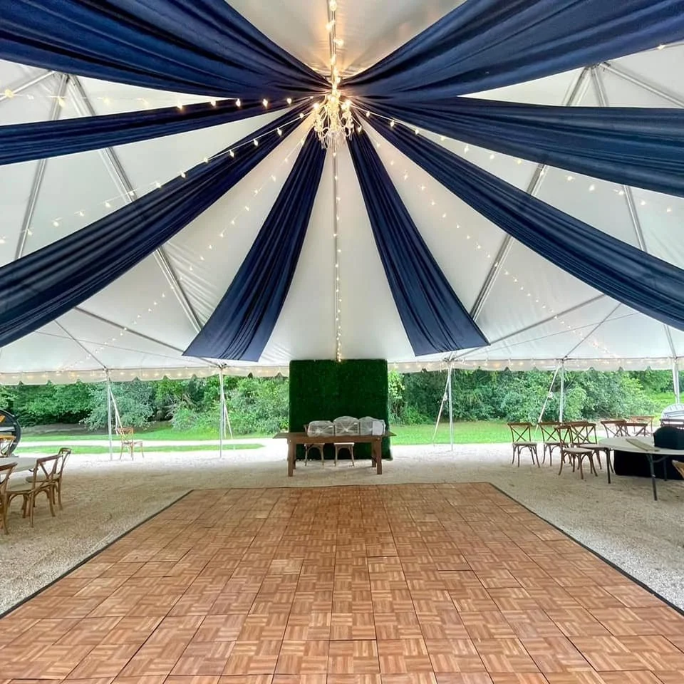 Inside a white event tent decorated with dark blue drapes and string lights. There is a checkered wooden dance floor and a table against a green backdrop. Chairs and additional tables are visible around the tent perimeter.