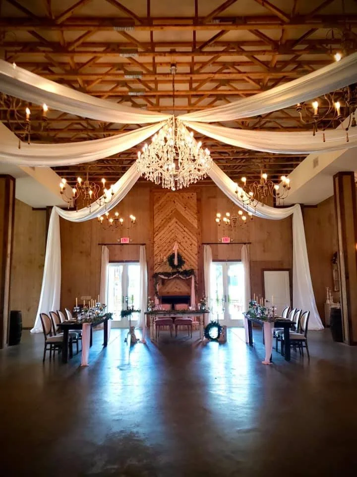 Elegantly decorated event hall with chandeliers, draped fabric on the ceiling, and a formal dining setup with a wooden backdrop.
