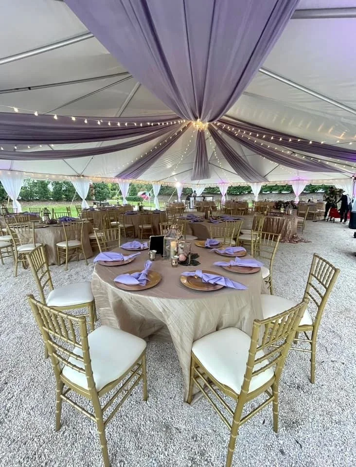 Outdoor tent with tables set for an event, featuring purple drapery, string lights, gold chairs, and round tables with beige tablecloths and lilac napkins.