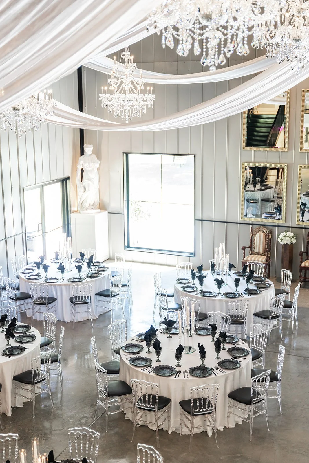 Elegant event venue with round tables, white tablecloths, black napkins, and clear chairs, under crystal chandeliers and draped fabric ceiling.
