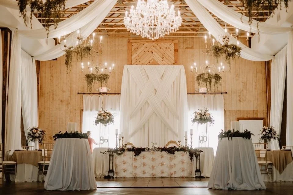 Elegant wedding reception venue with draped white fabric, chandeliers, floral arrangements, and decorated tables.