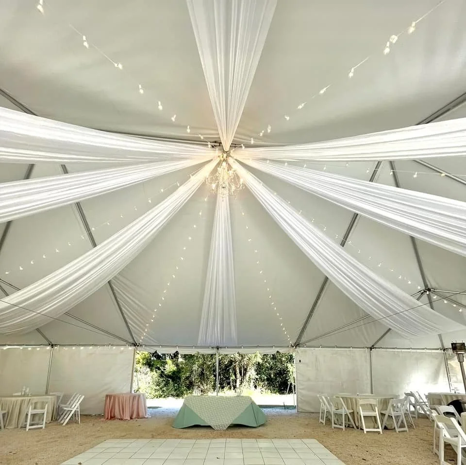 Interior of a large event tent with white drapes on the ceiling, string lights, and arranged tables and chairs. A chandelier hangs in the center.