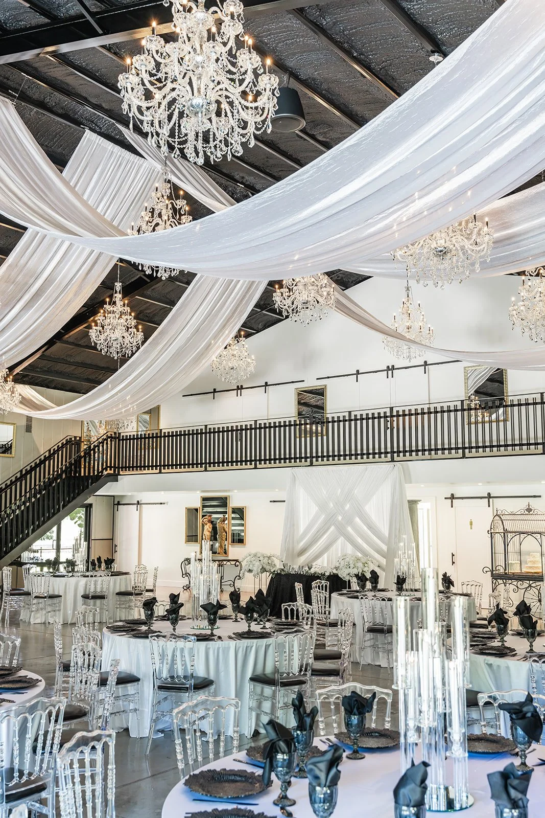 Elegantly decorated event space with chandeliers, draped fabric ceiling, round tables set with black and white linens, transparent chairs, black napkins on plates, and decorative glassware.