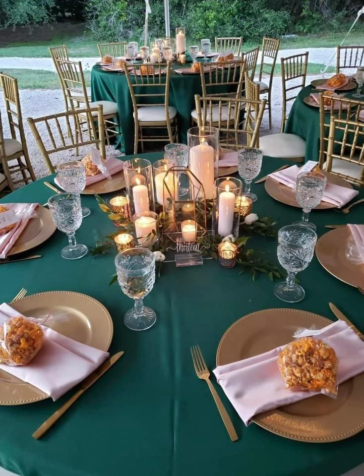 Elegant table setting with green tablecloth, gold chairs and plates, glassware, candles, and floral centerpiece outdoors.