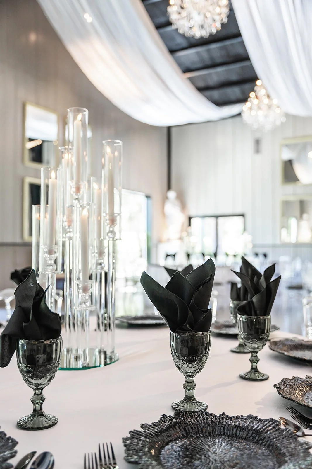 Elegant dining setup with black napkins in glasses, textured plates, and tall candlesticks, under draped white fabric and chandeliers.