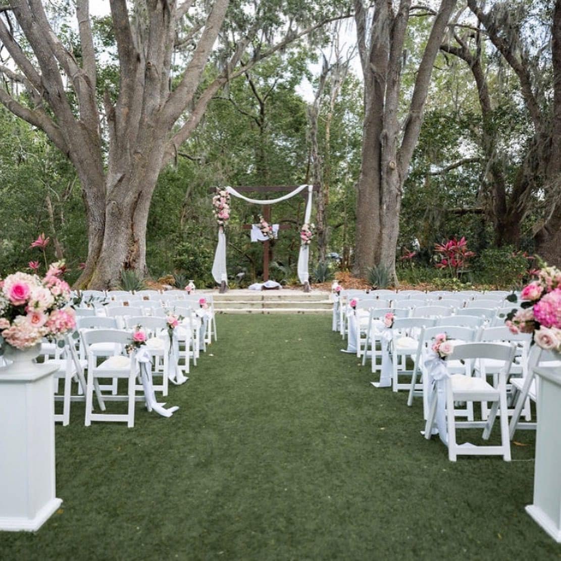 Outdoor wedding ceremony setup with white chairs and floral decorations under large trees.