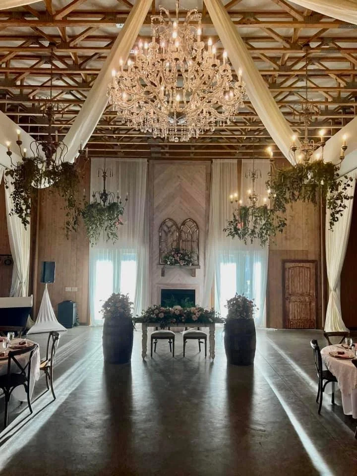 Elegant event venue with a large chandelier, draped ceilings, floral arrangements, and rustic decor.