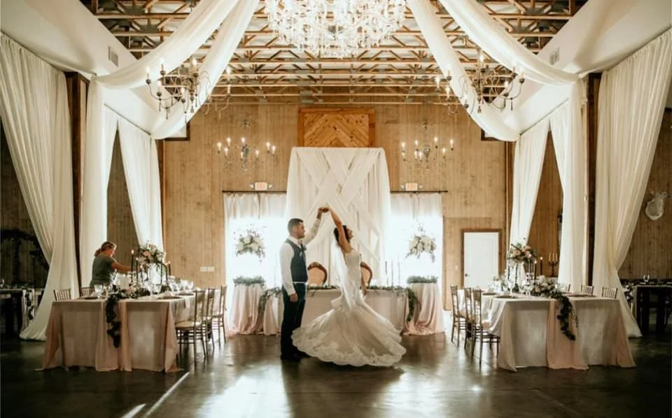 A couple is dancing in a beautifully decorated wedding venue with draped white curtains and chandeliers. The room is elegantly set with tables adorned with floral arrangements and a soft pink color theme.