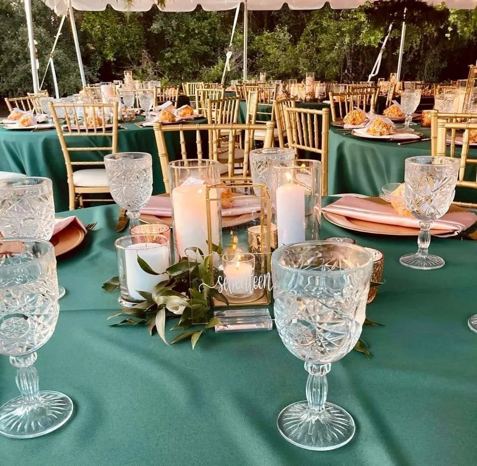 Elegant outdoor wedding reception setup with green tablecloths, crystal glasses, gold chairs, and candles; place settings include decorative foliage.