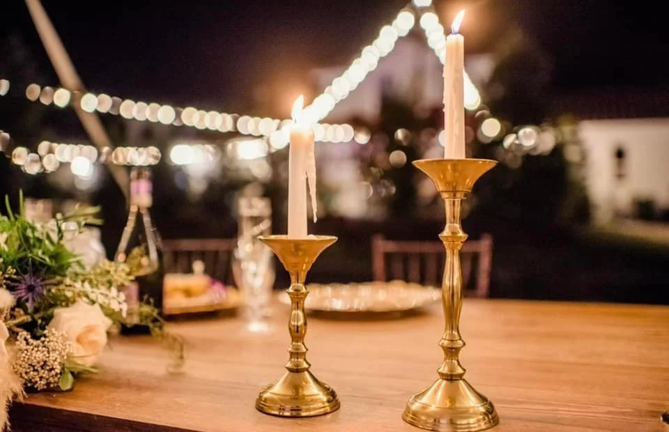 Two lit candles in brass holders on a wooden table, with string lights blurred in the background.
