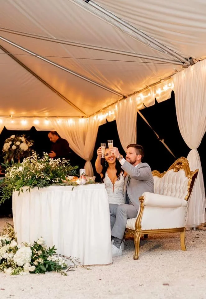 A bride and groom sitting at a decorated table under a tent, toasting with champagne glasses. The setting includes white drapes, string lights, and floral arrangements.