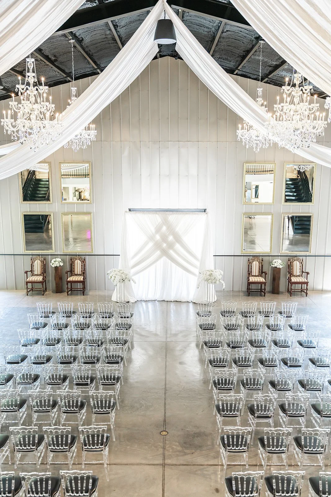 Elegant indoor wedding venue with a white draped altar, clear chairs, chandeliers, and decorative mirrors.