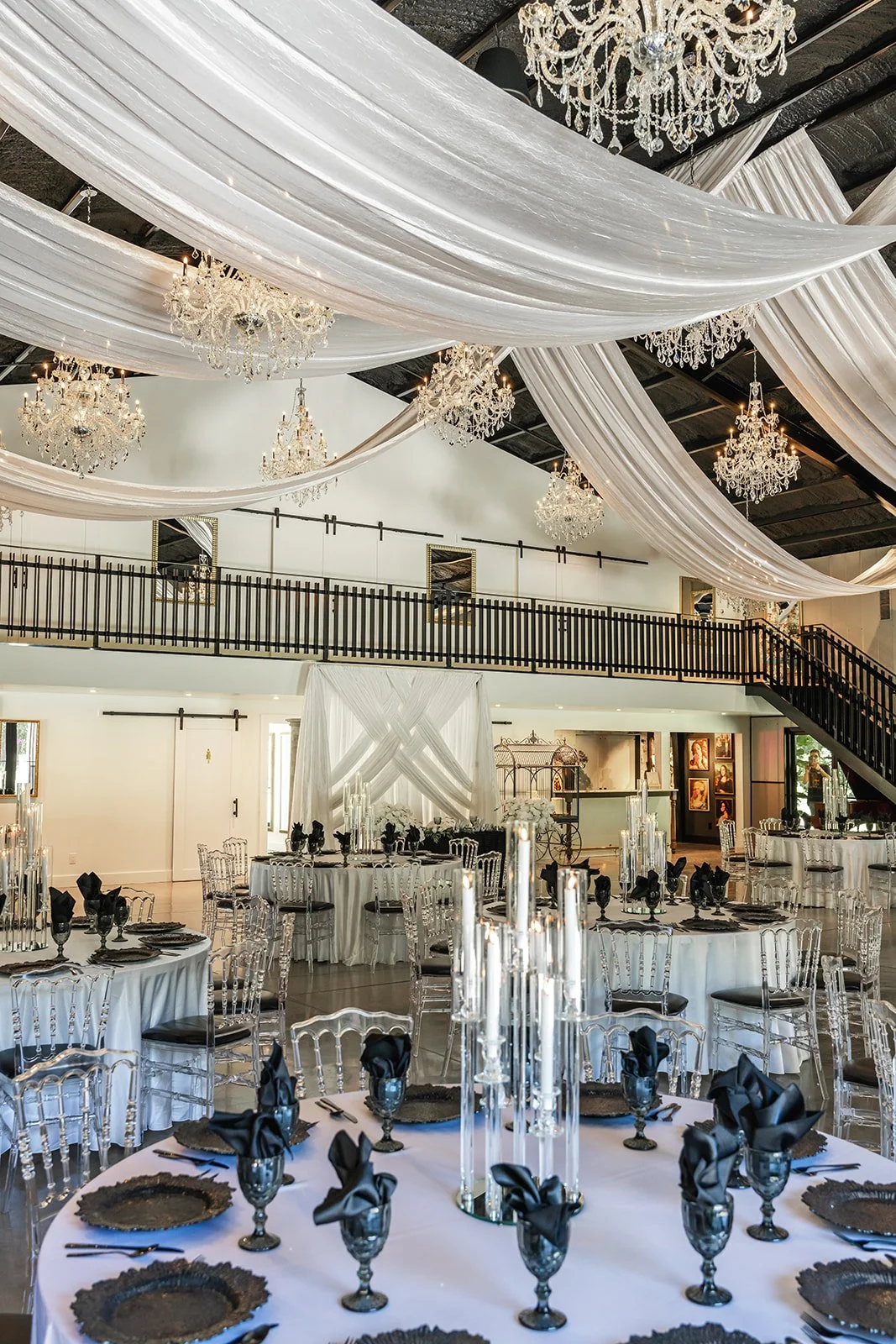 Elegant event hall with white draped ceilings, chandeliers, and set tables with black and white tableware.