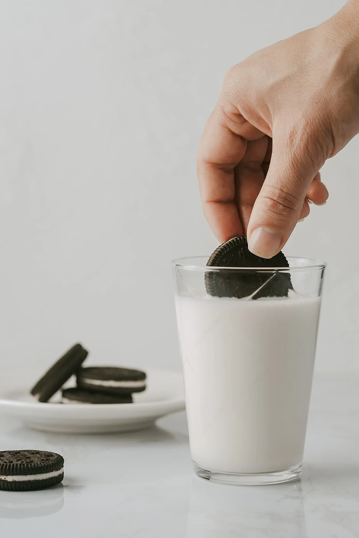 a hand dunking an oreo in a glass of milk with a plate of oreos next to it