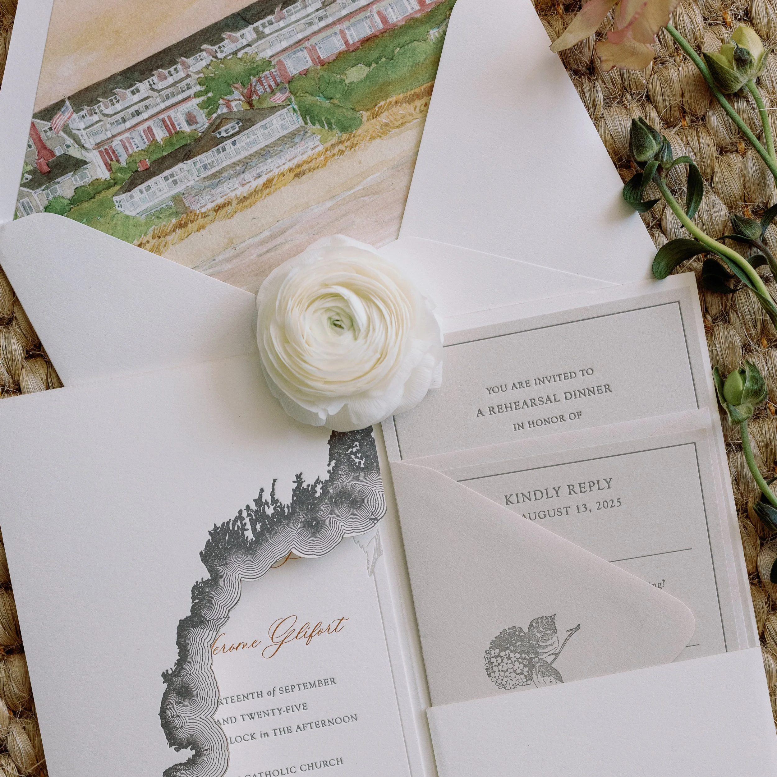 Invitation card with floral and artistic details, wedding details, and reply card, surrounded by a white flower, greenery, and an illustrated card of a waterfront house, laid on a textured woven surface.