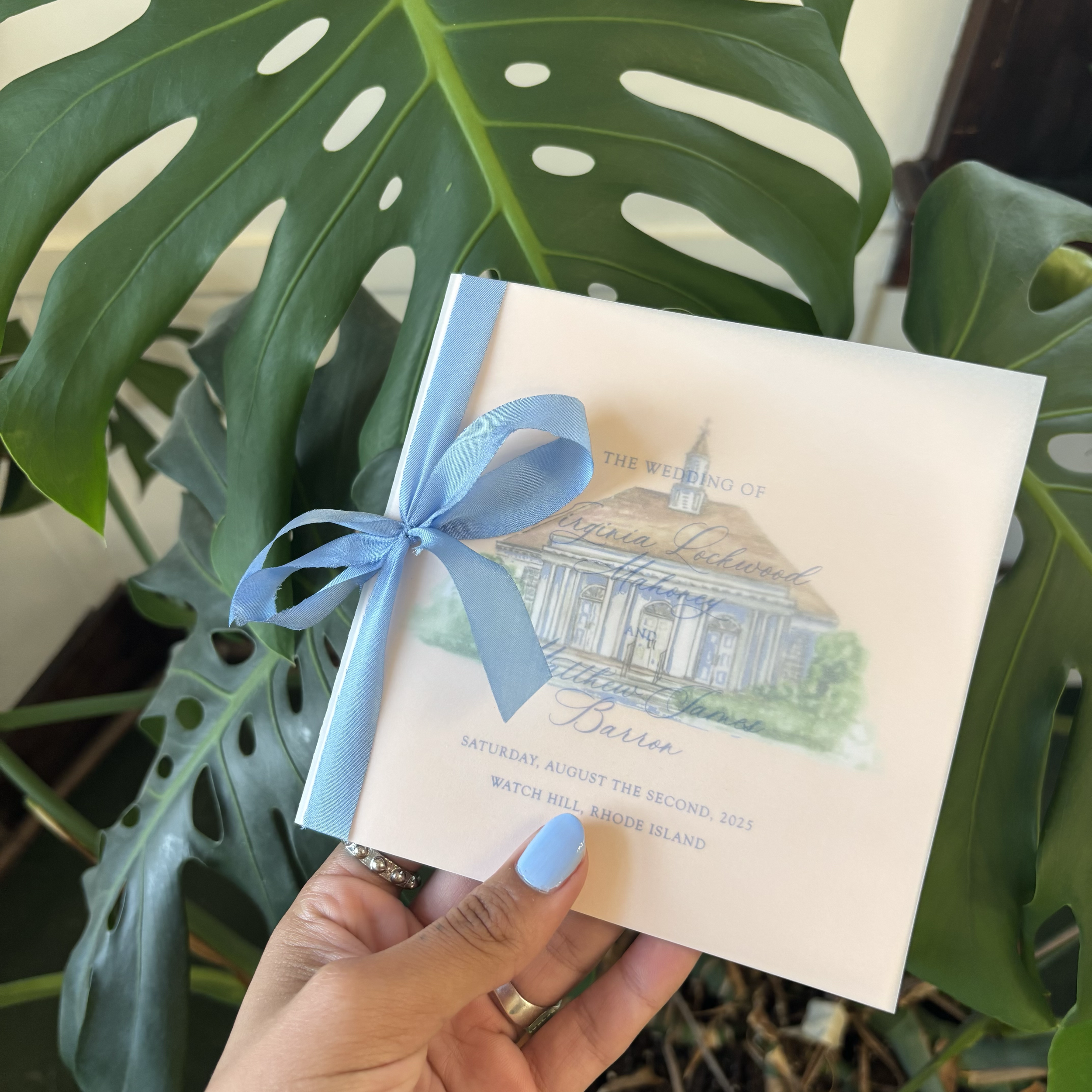 A wedding invitation with a blue ribbon bow, held in front of a large green monstera plant. The invitation features an illustration of a house and expects guests to attend on Saturday, August 2, 2025, in Rhode Island.