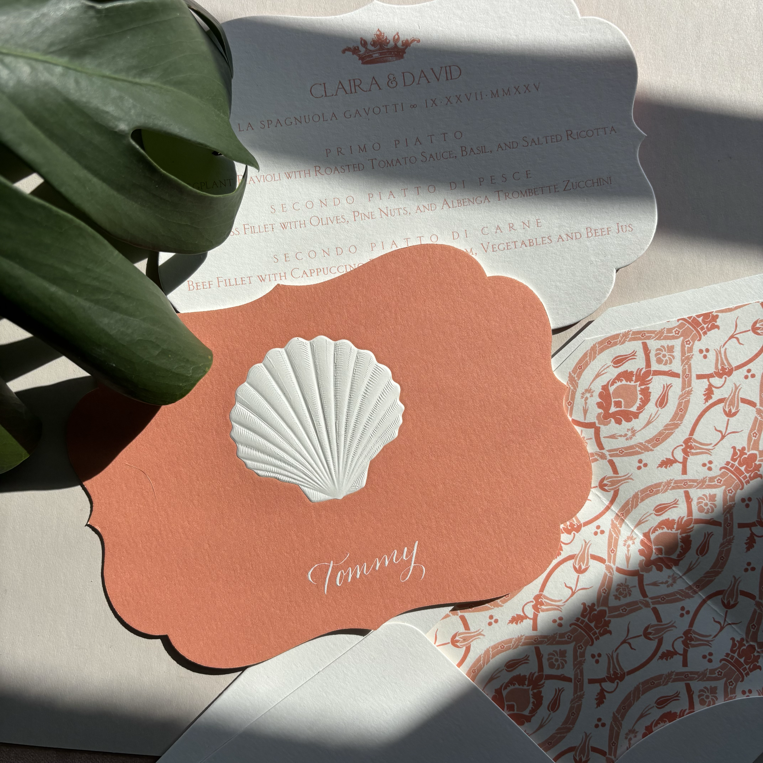 A wedding invitation with a coral-colored scalloped edge and a white embossed shell design, with the name "Tommy" written in cursive. The invitation is partially covered by some green leaves and other decorative paper elements, with one sheet showing