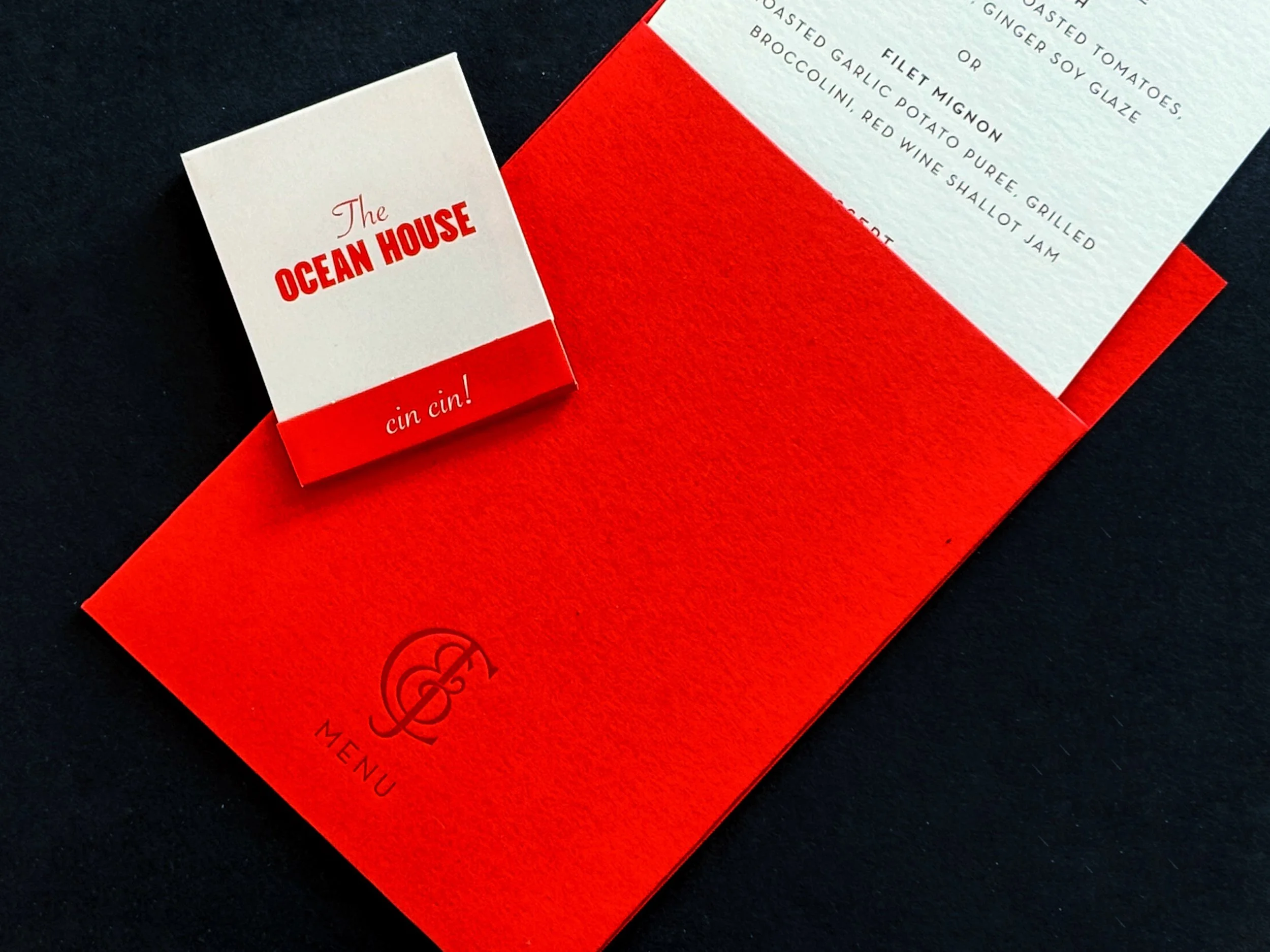 A red and white restaurant menu and a small card on a black background. The menu has a logo with the letter 'T' and the word 'MENU' on it. The small card reads 'The Ocean House' with a red band underneath that says 'cin cin !'