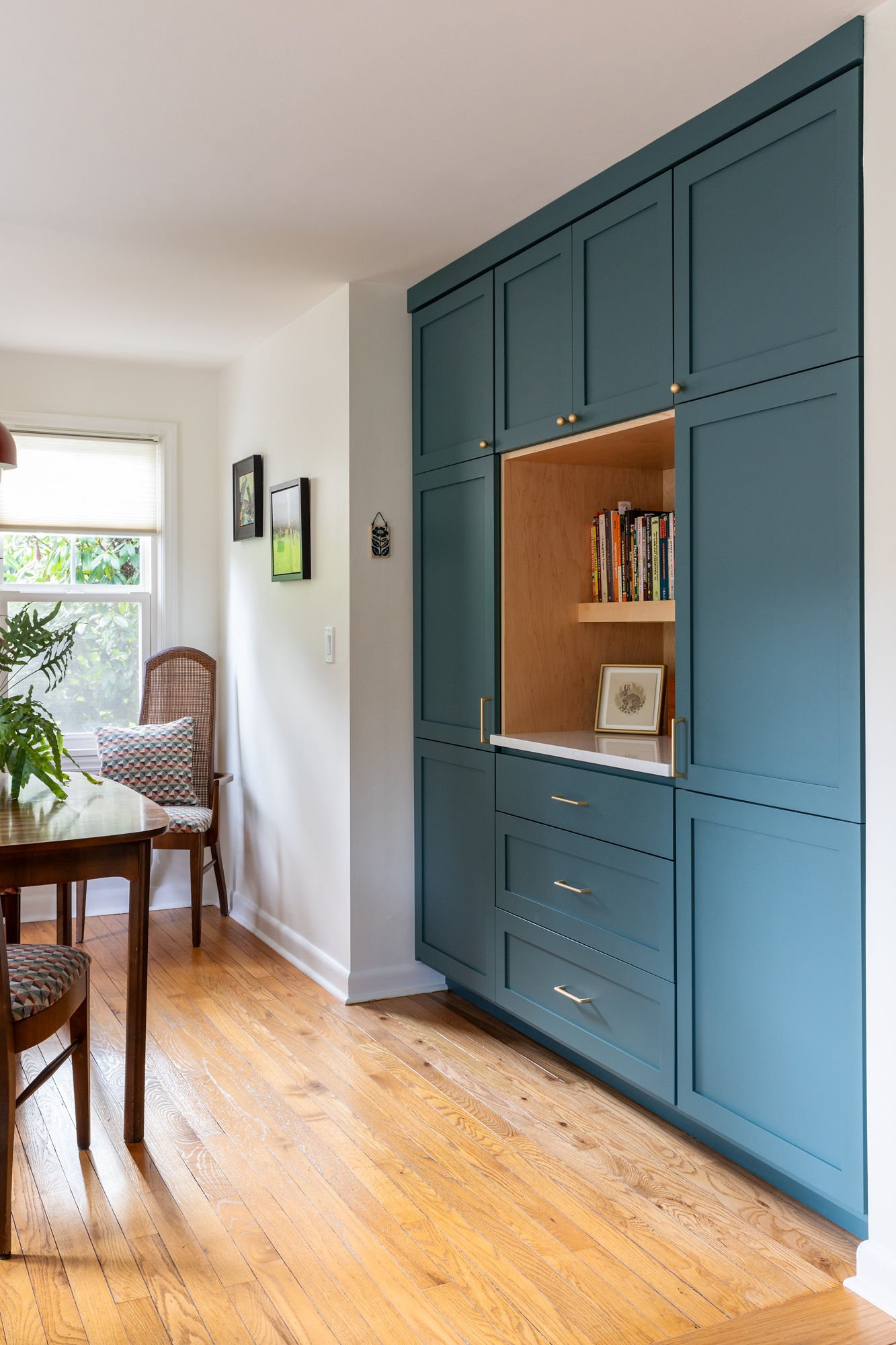 A blue built-in cabinet with gold handles and open wooden shelves, holding books and framed pictures, in a bright room with hardwood flooring and a window.