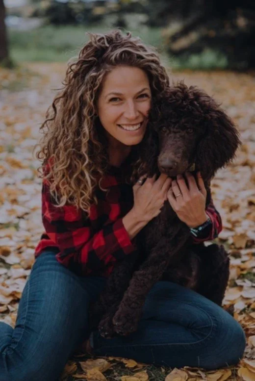 A woman with curly hair smiling and holding a large black dog sitting on autumn leaves.