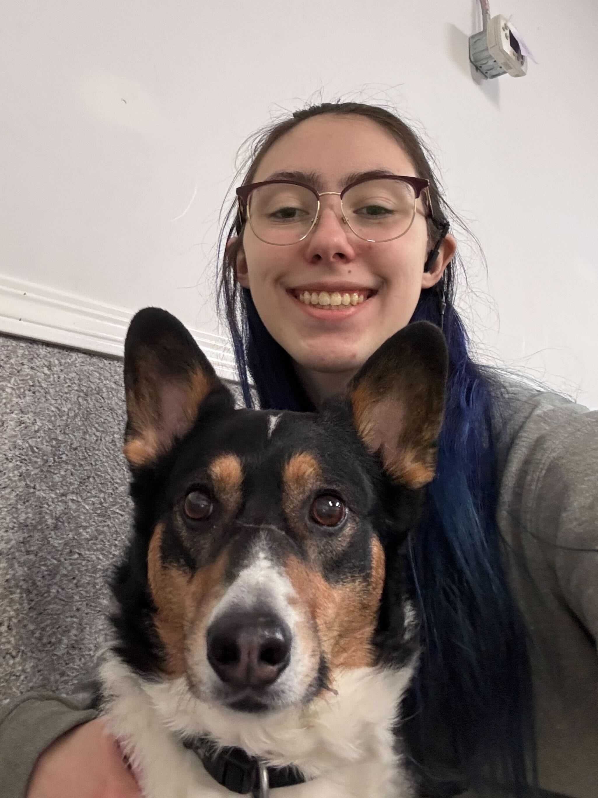 A smiling person with glasses and blue hair taking a selfie with a black and tan dog with large ears.