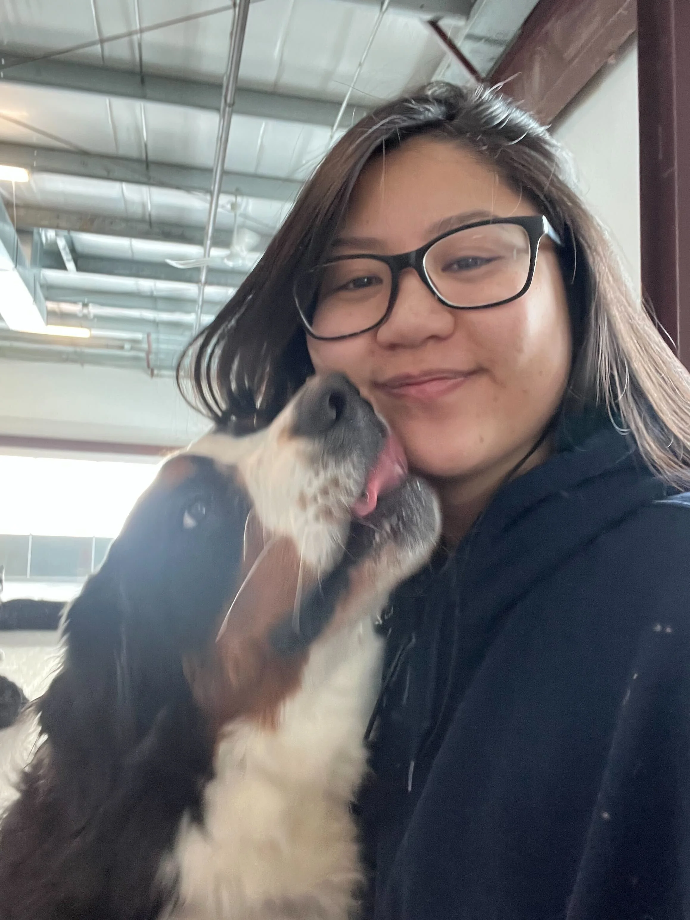 A woman with glasses smiling as a dog licks her face inside a building with metal ceiling and large window in Airdrie Alberta.