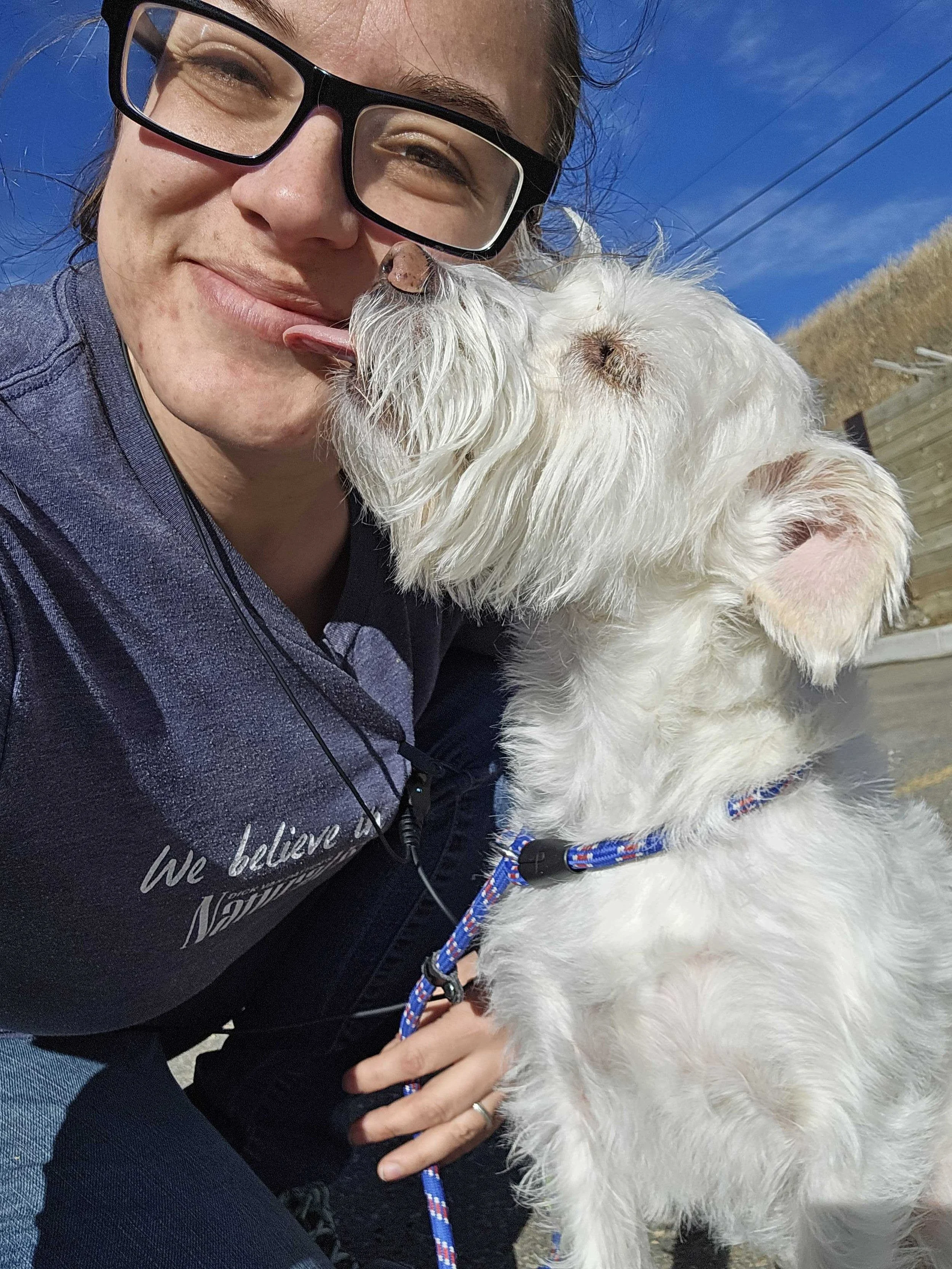 Holly a Daycare team leader is outdoors, smiling as a white dog licks her face. The dog has a colorful collar and leash, and the background shows a wooden fence, blue sky, and dry grass in Airdrie Alberta