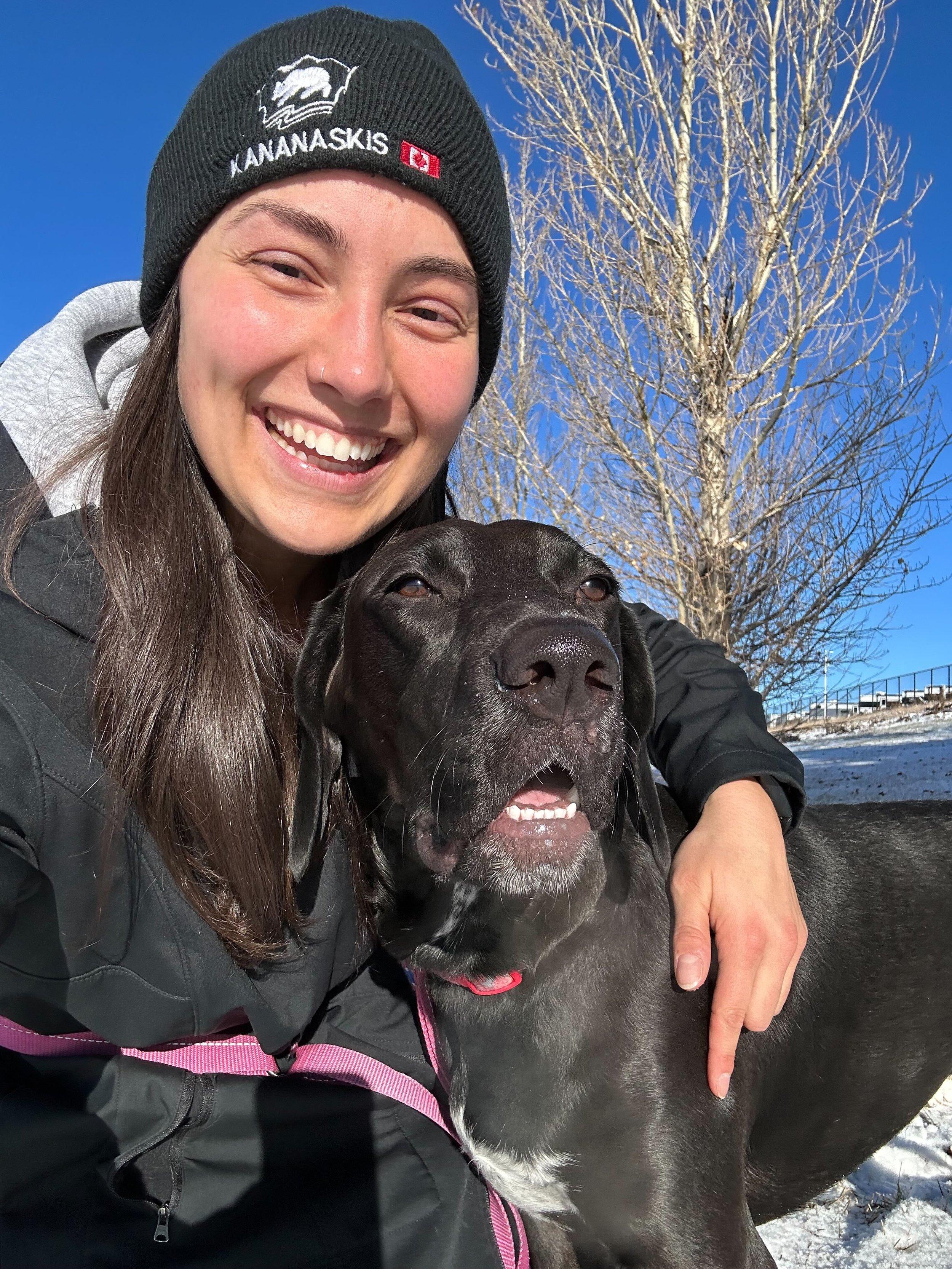 A smiling woman with long brown hair wearing a black Beanie hat with a logo and a gray hoodie, hugging a large black dog with white markings, against a snowy outdoor background with a clear blue sky and a leafless tree in Airdrie, Alberta, Canada