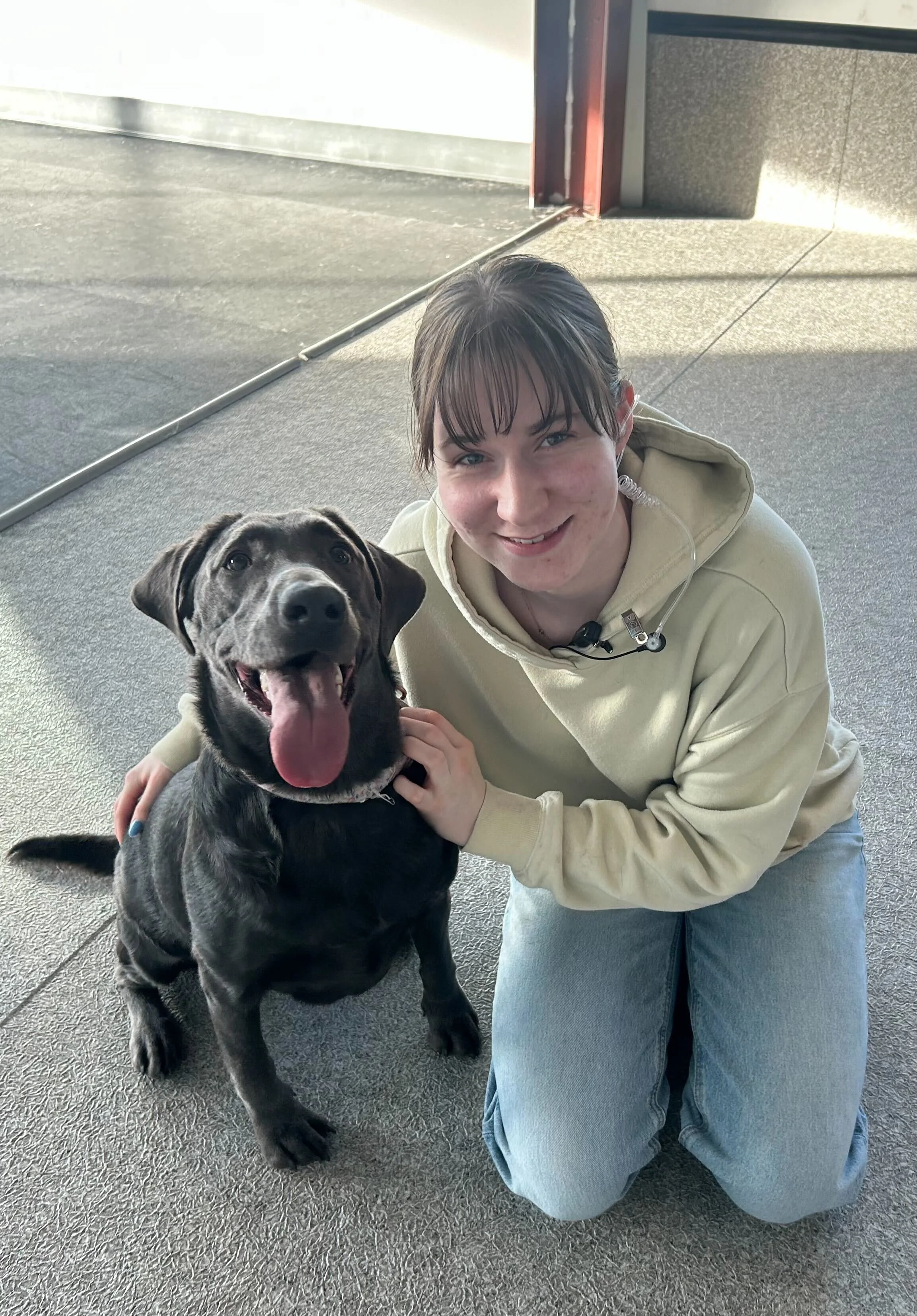 A woman kneeling on the floor with a smile, holding and petting a black dog with a white patch on its chest. The dog is sitting with its tongue out. The setting appears to be indoors, with sunlight coming in at Airdrie Puppy Pals Dog Daycare.