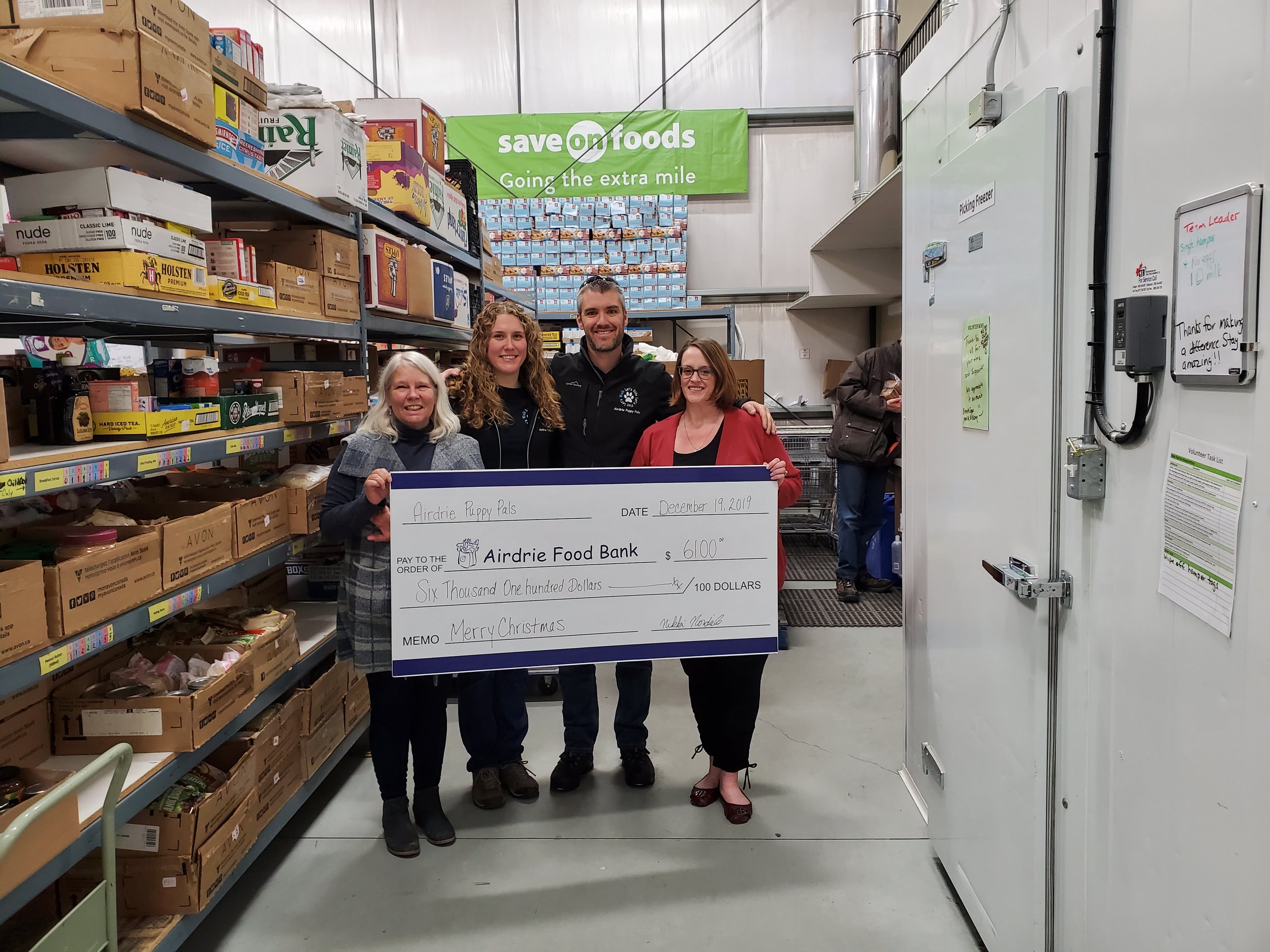 Four people standing inside a food bank holding an oversized check for $6100 made out to Airdrie Food Bank, dated December 19, 2019, with a Christmas message. They are smiling, surrounded by shelves of food items and a banner that says "save on foods" in the background.