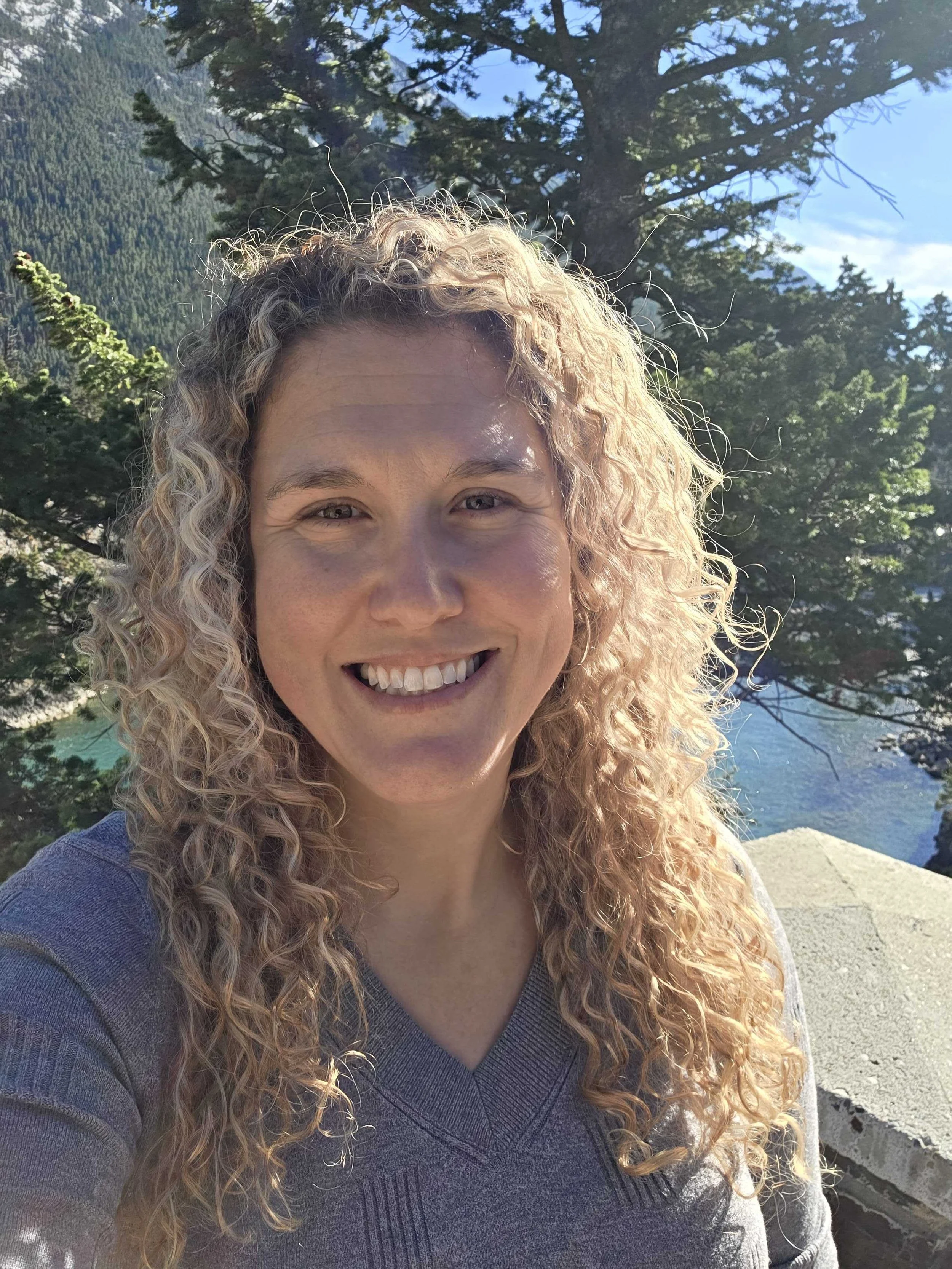 A woman with curly blonde hair smiling outdoors with trees and a river in the background.