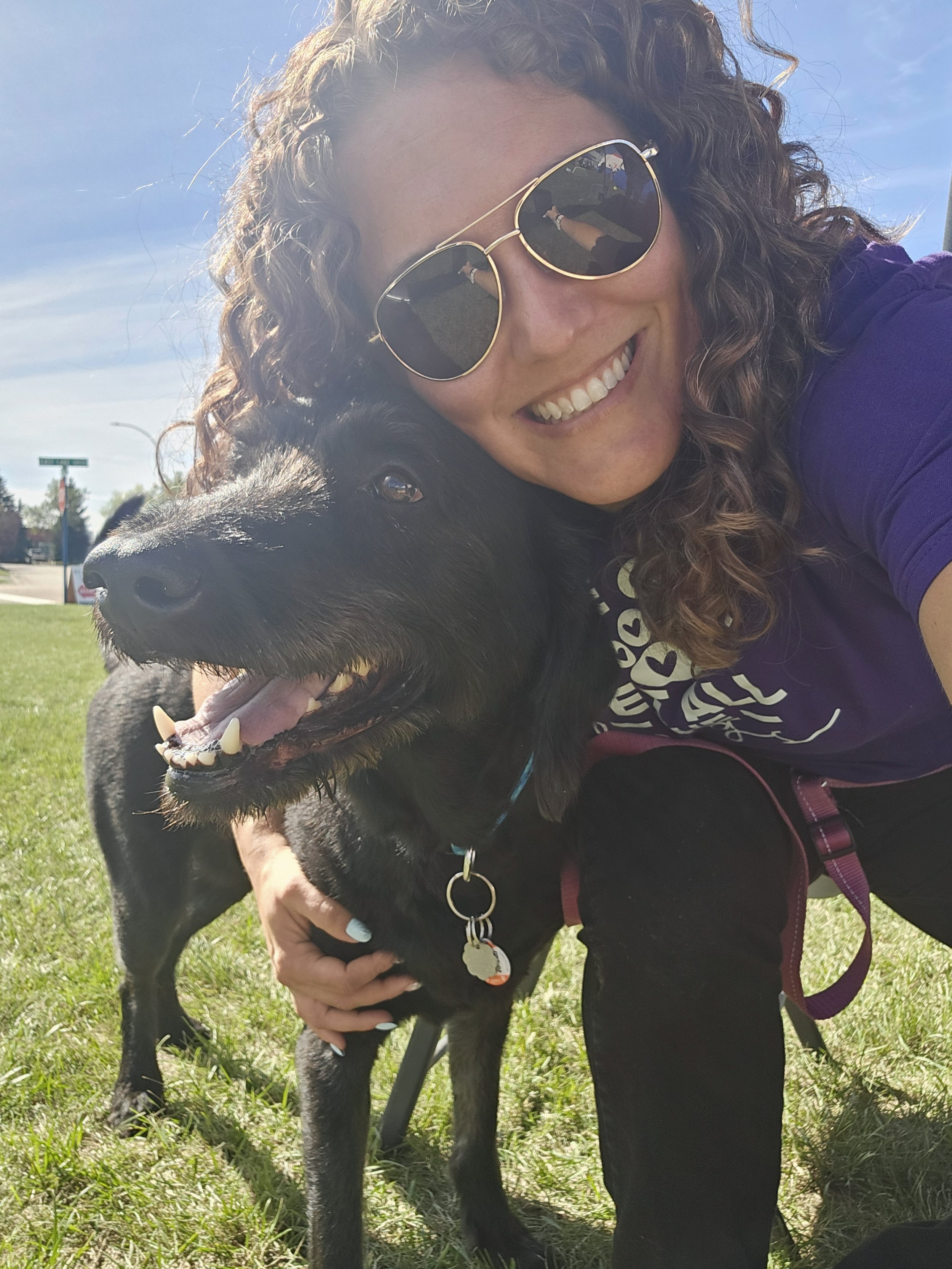 Nikki Nordick A woman with curly hair, wearing sunglasses, smiling and hugging a black dog with an open mouth and tongue out, outdoors on a sunny day.  Nikki is a Certified Dog Behavior Consultant