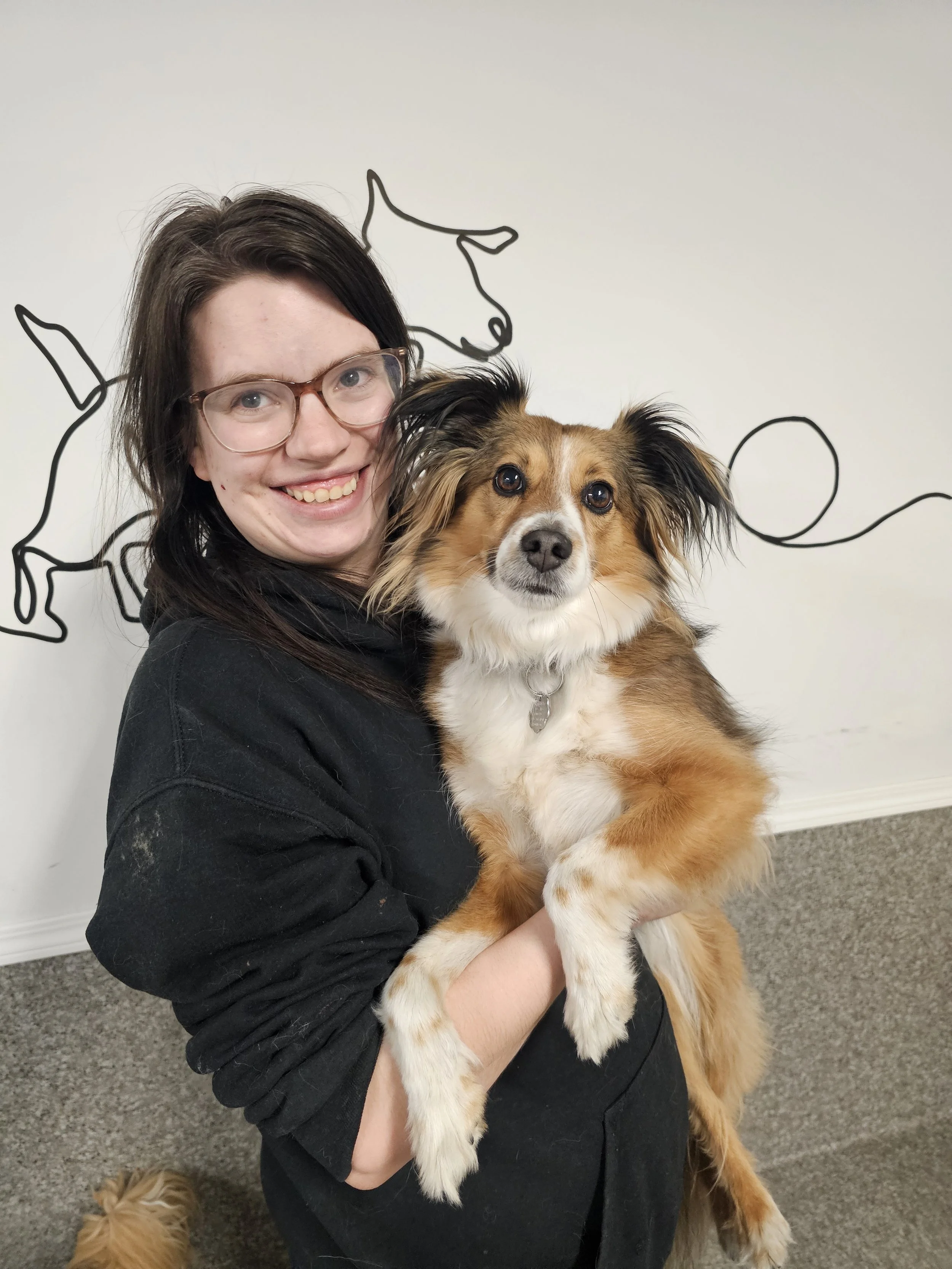 A woman with glasses and dark hair smiling while holding a medium-sized dog with a tri-color coat and black ears. There is a line art drawing of dogs on the wall behind them in Airdrie, Alberta.