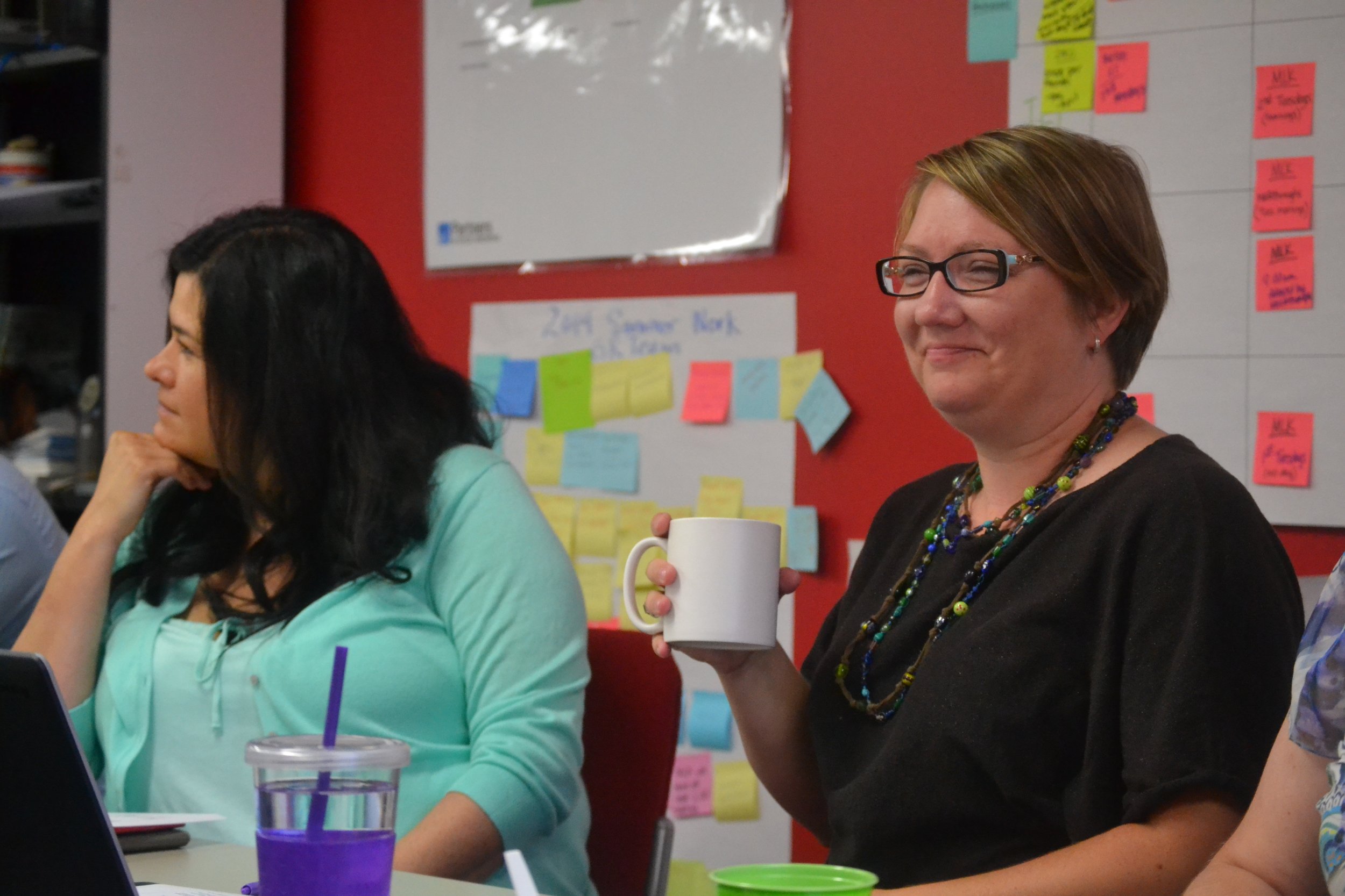 Photo of two women smiling and seated at a table, paying attention to an off-screen speaker. Behind them is a colorful wall with brainstorming notes.