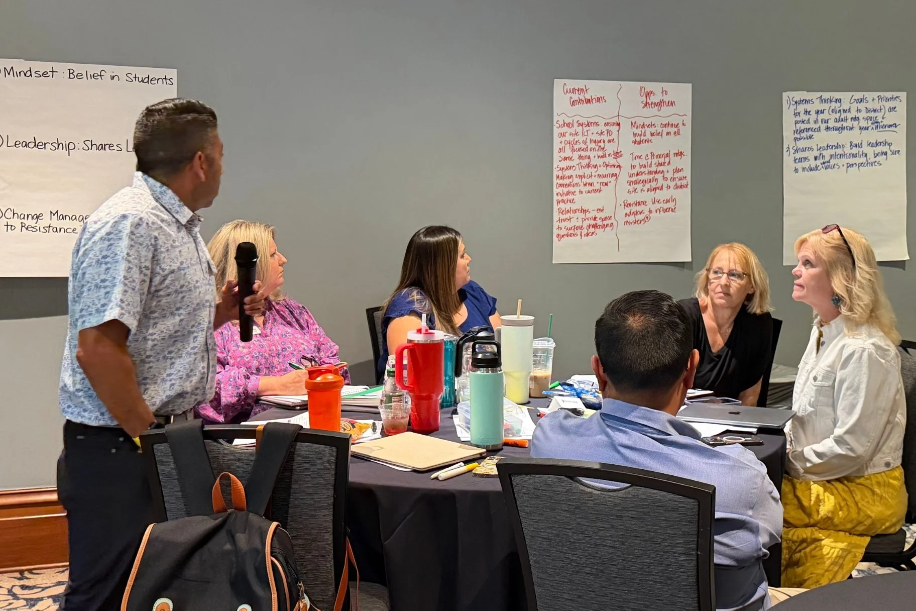 Photo of a person with a microphone explaining what is written on a document posted on the wall of a conference room. People seated around a table are listening intently.