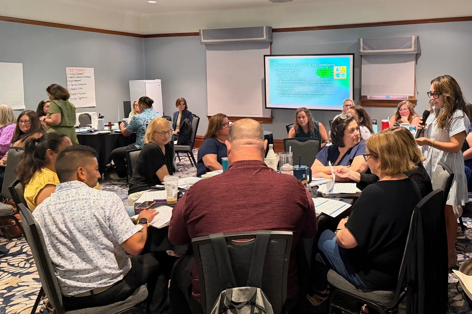 Photo of a meeting room full of round tables where school and district leaders are seated, listening to a person with a microphone. A projector screen is in the background.