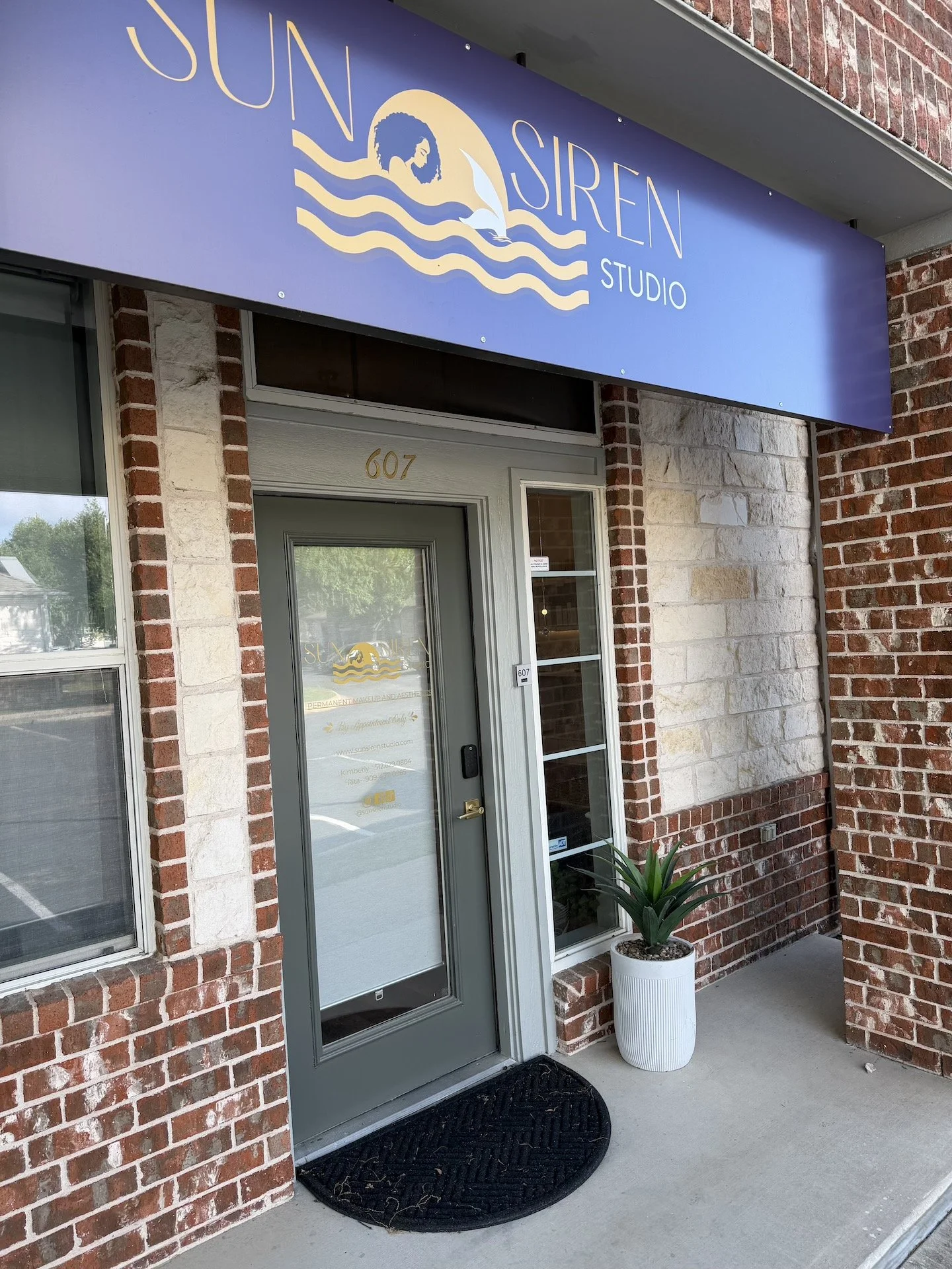Storefront with a blue sign reading 'Sun Siren Studio' featuring a woman with curly hair, waves, and a dolphin logo. The entrance has a gray door with glass panels, a black rug in front, and a potted plant beside the door.