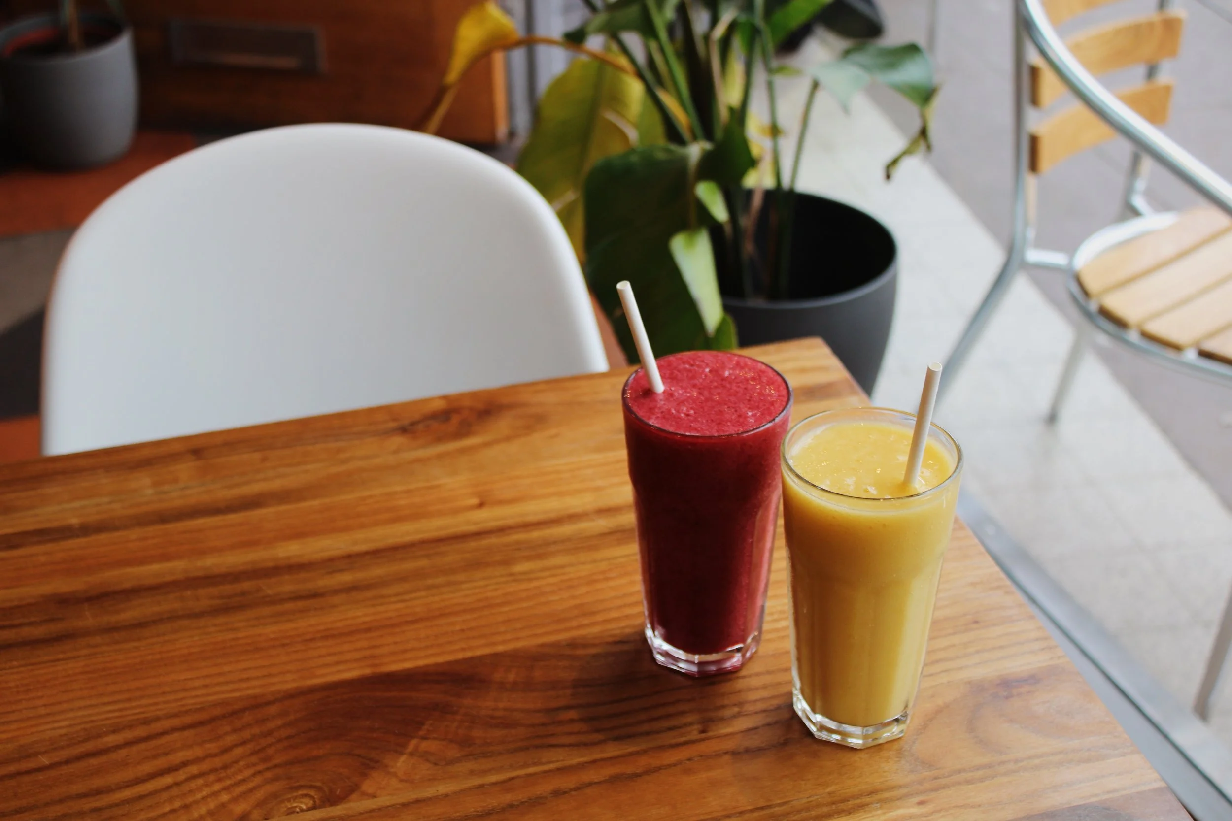 Two tall glasses of smoothies on a wooden table, one red and one yellow, with white straws. There is a white chair, a potted plant, and outdoor seating in the background.