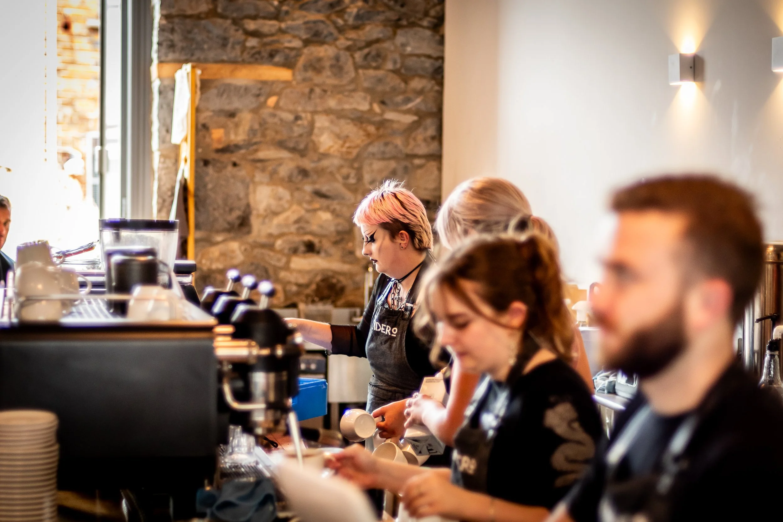 Baristas working behind a coffee counter in a cafe, with a stone wall in the background, and a window letting in natural light.