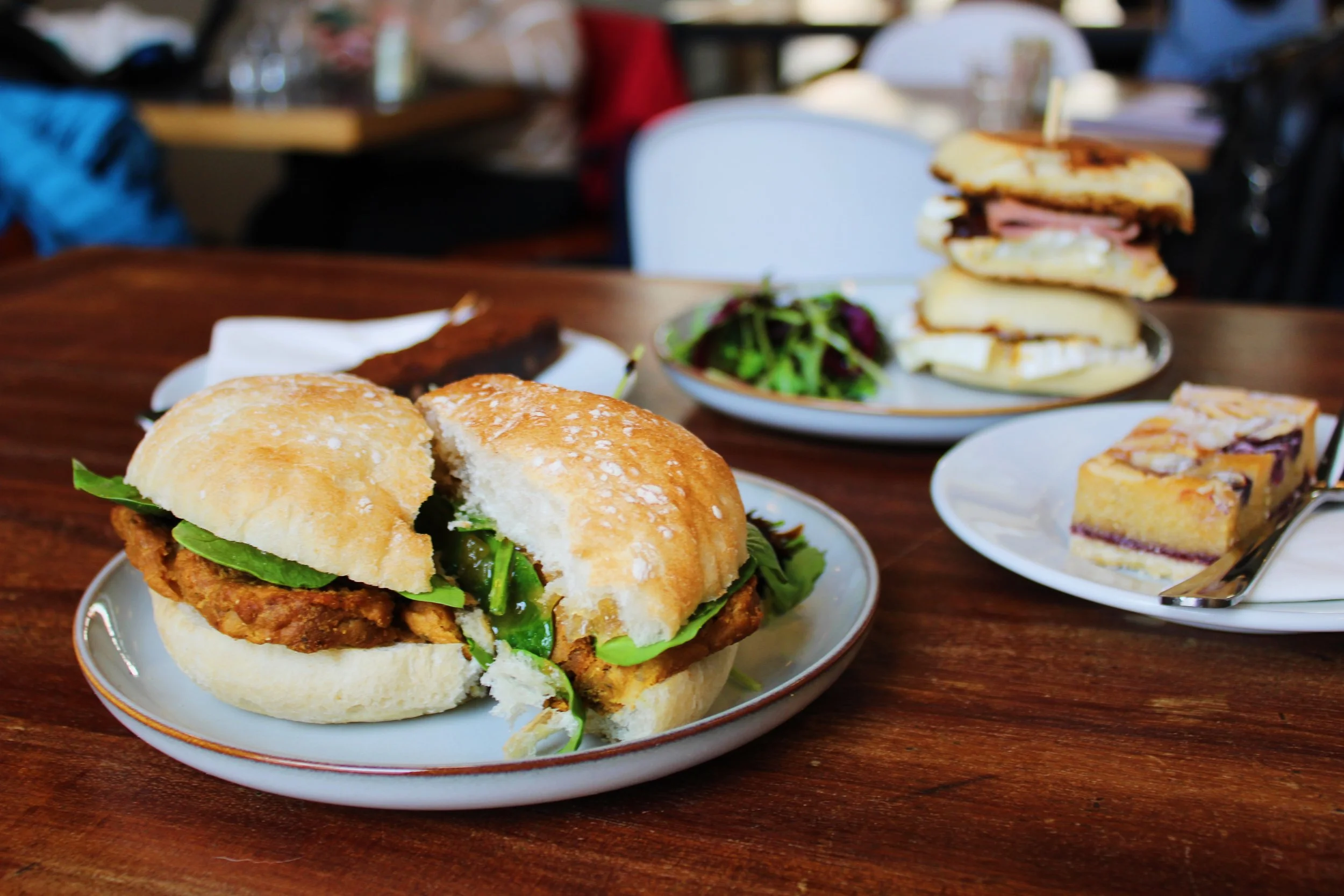A plate with a sandwich containing falafel, lettuce, and pickles on a bun, a side of greens, and another plate with sandwiches and cake slices on a wooden table in a restaurant setting.