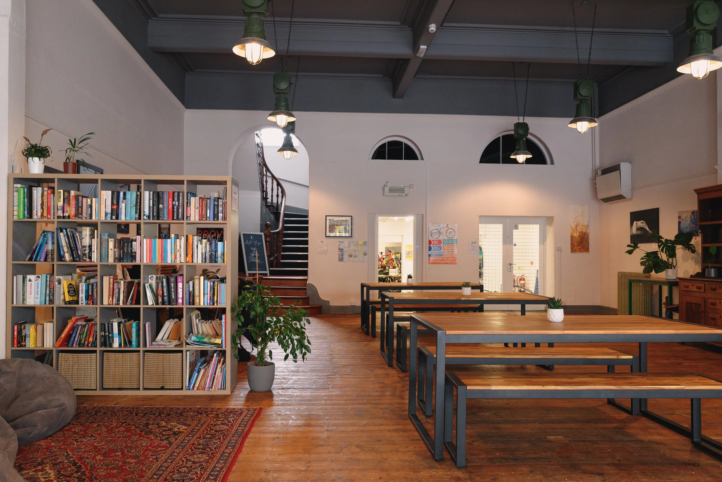 Coffee shop interior with wooden tables, a bookshelf filled with books, indoor plants, hanging light fixtures, and a staircase leading to the upper level.