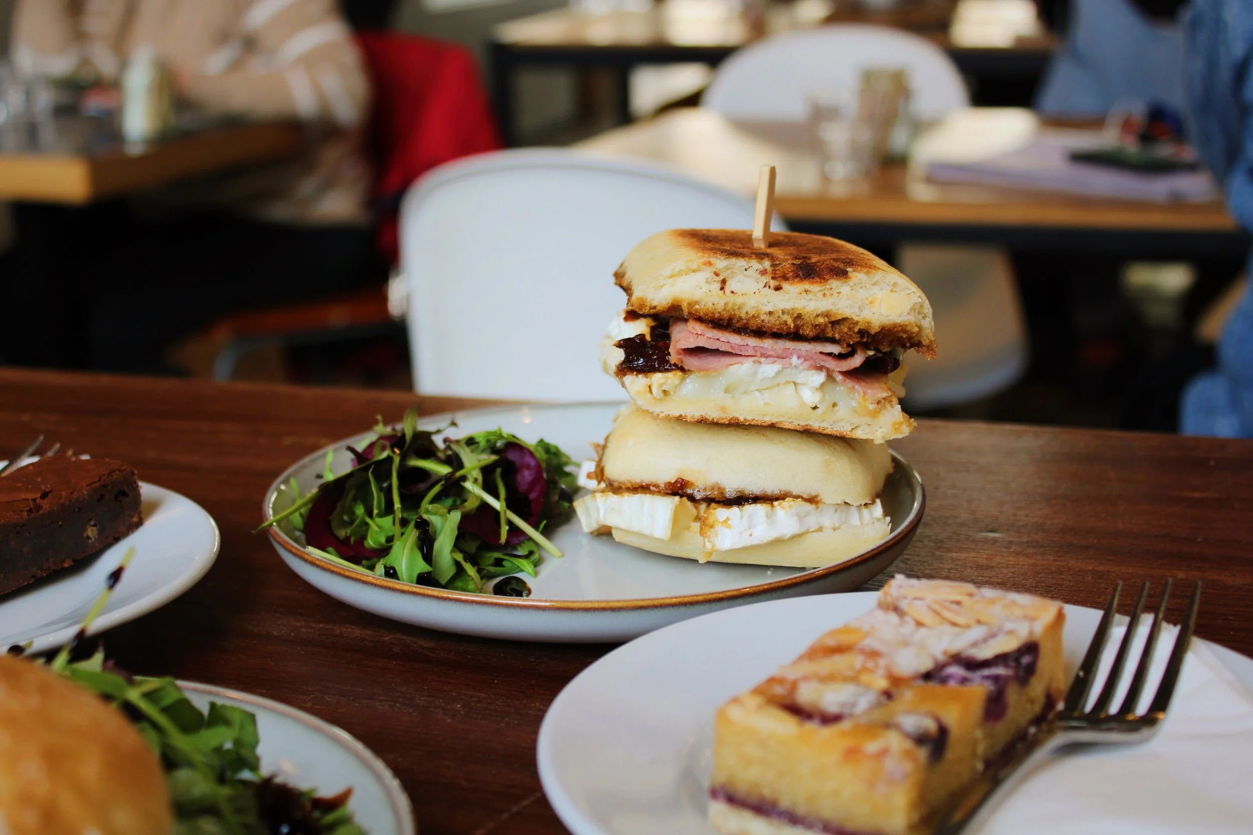 A stack of three sandwiches on a plate, accompanied by a side salad, on a wooden table in a restaurant.