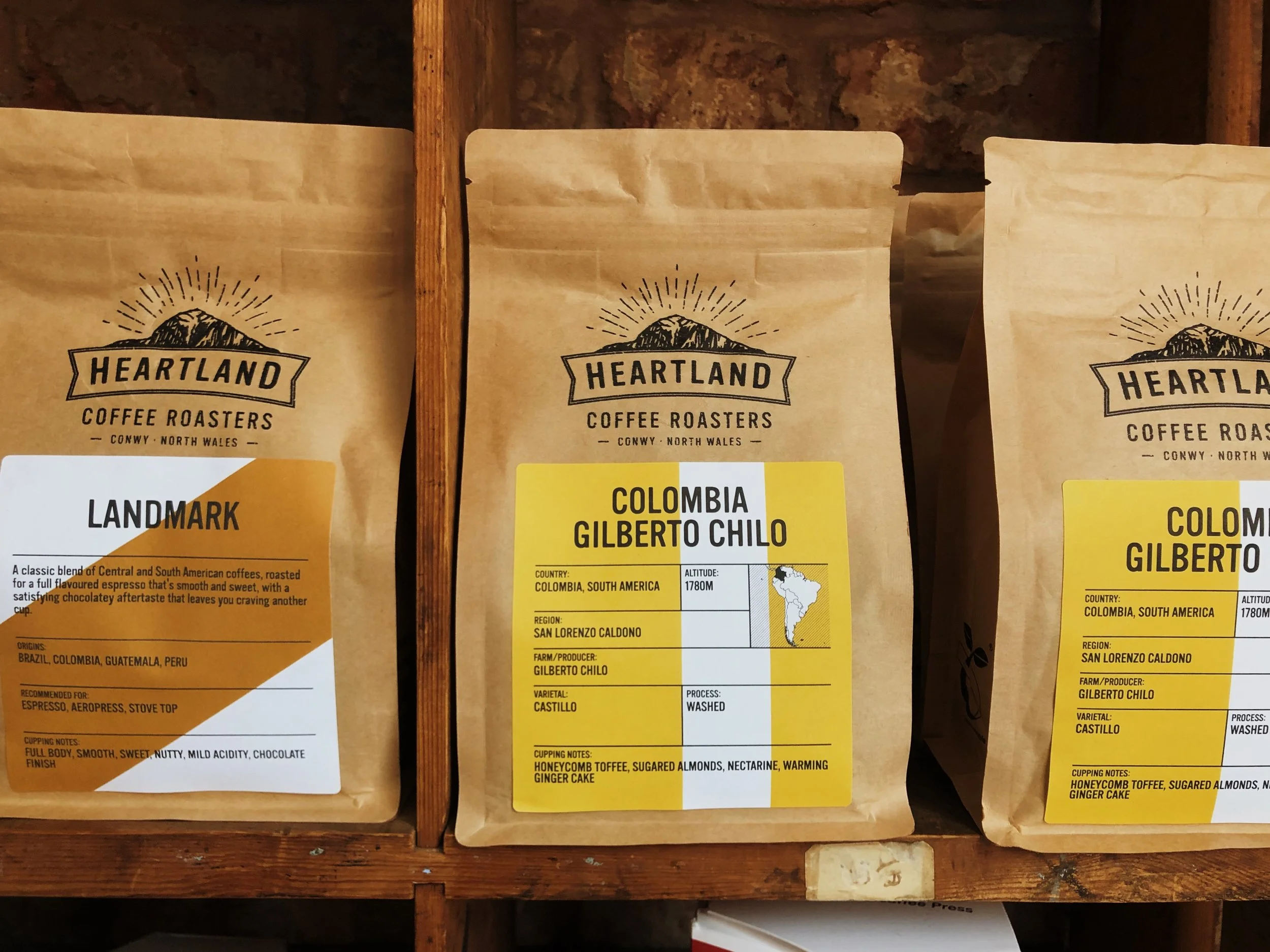 Three brown paper bags of coffee from Heartland Coffee Roasters, with the middle bag labeled 'Colombia Gilberto Chilo' and the left bag labeled 'Landmark'.