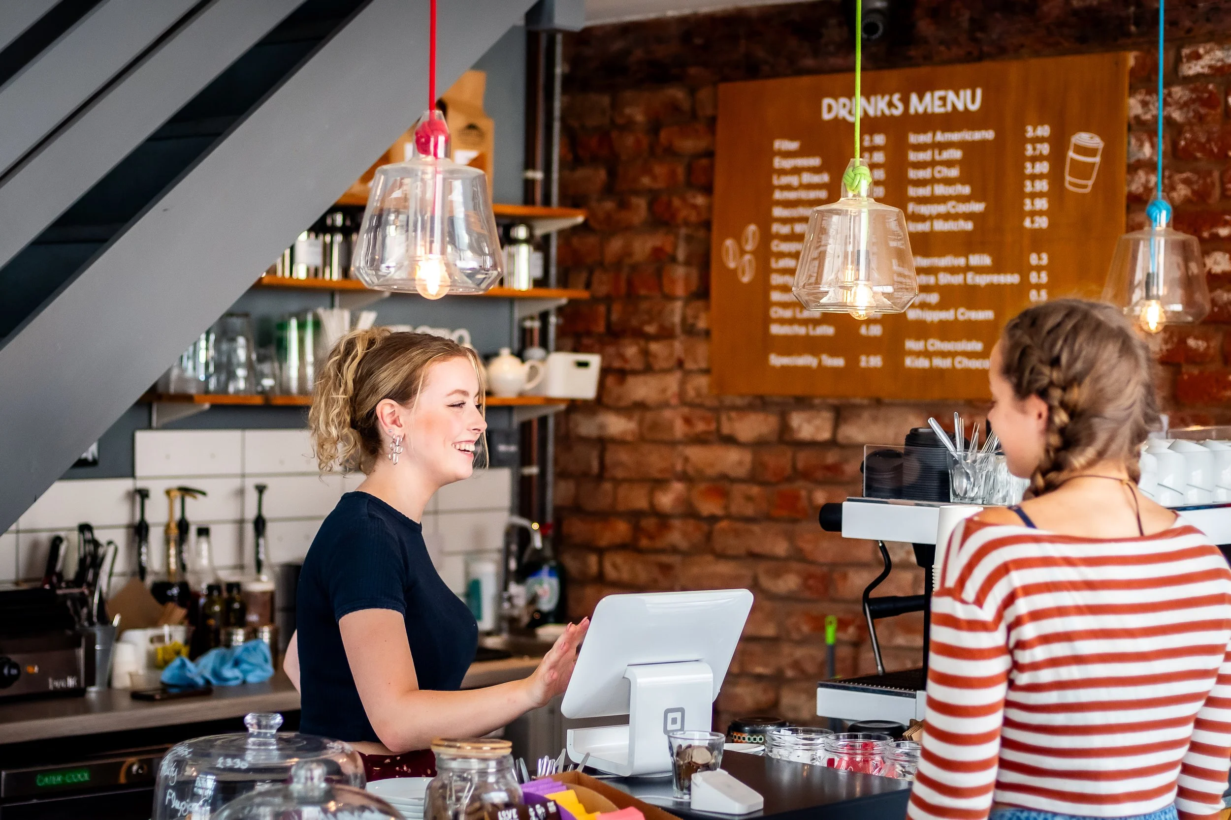 A woman working as a barista serving a customer at a coffee shop, with a drink menu on a brick wall in the background and pendant lights hanging from the ceiling.