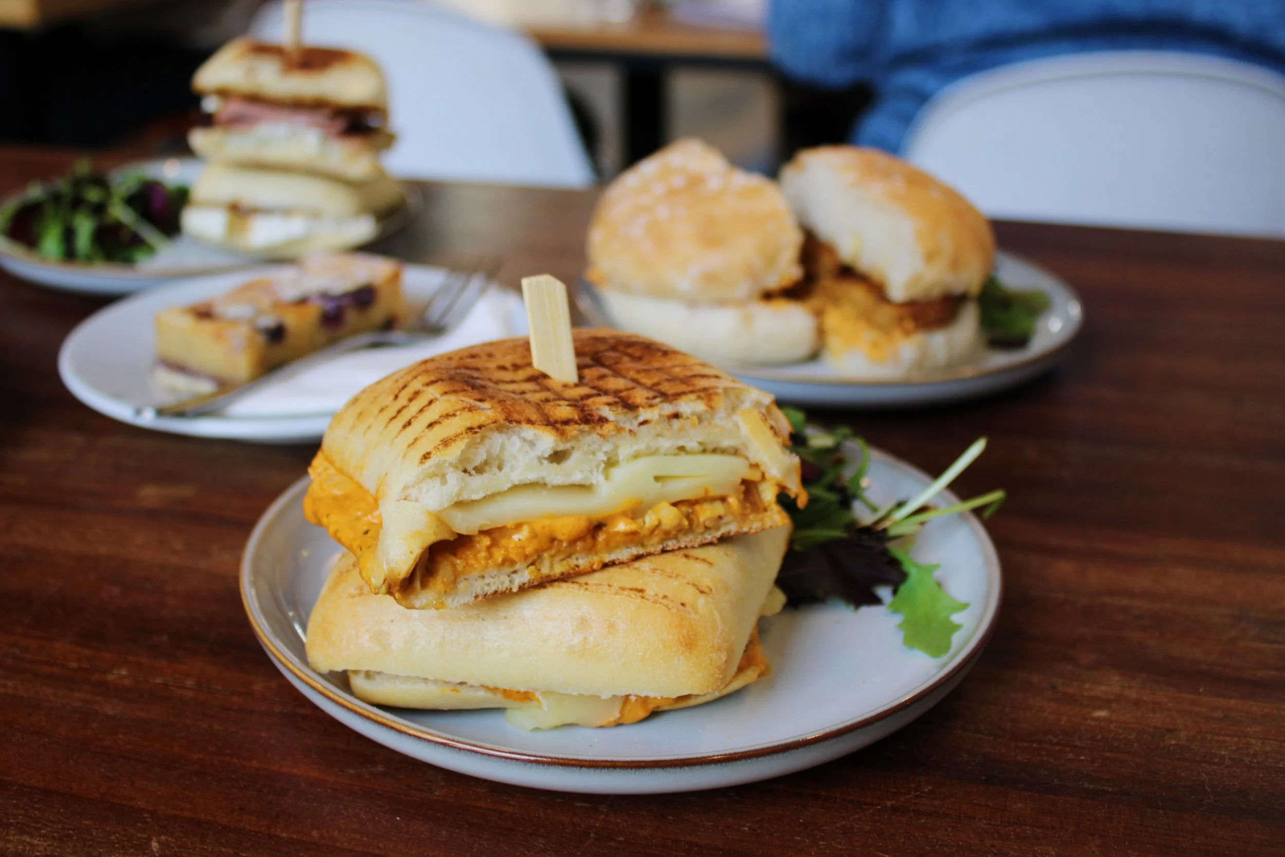 Close-up of a grilled sandwich with cheese, chicken, and sauce on a small white plate, with other sandwiches and a slice of cake in the background on a wooden table.