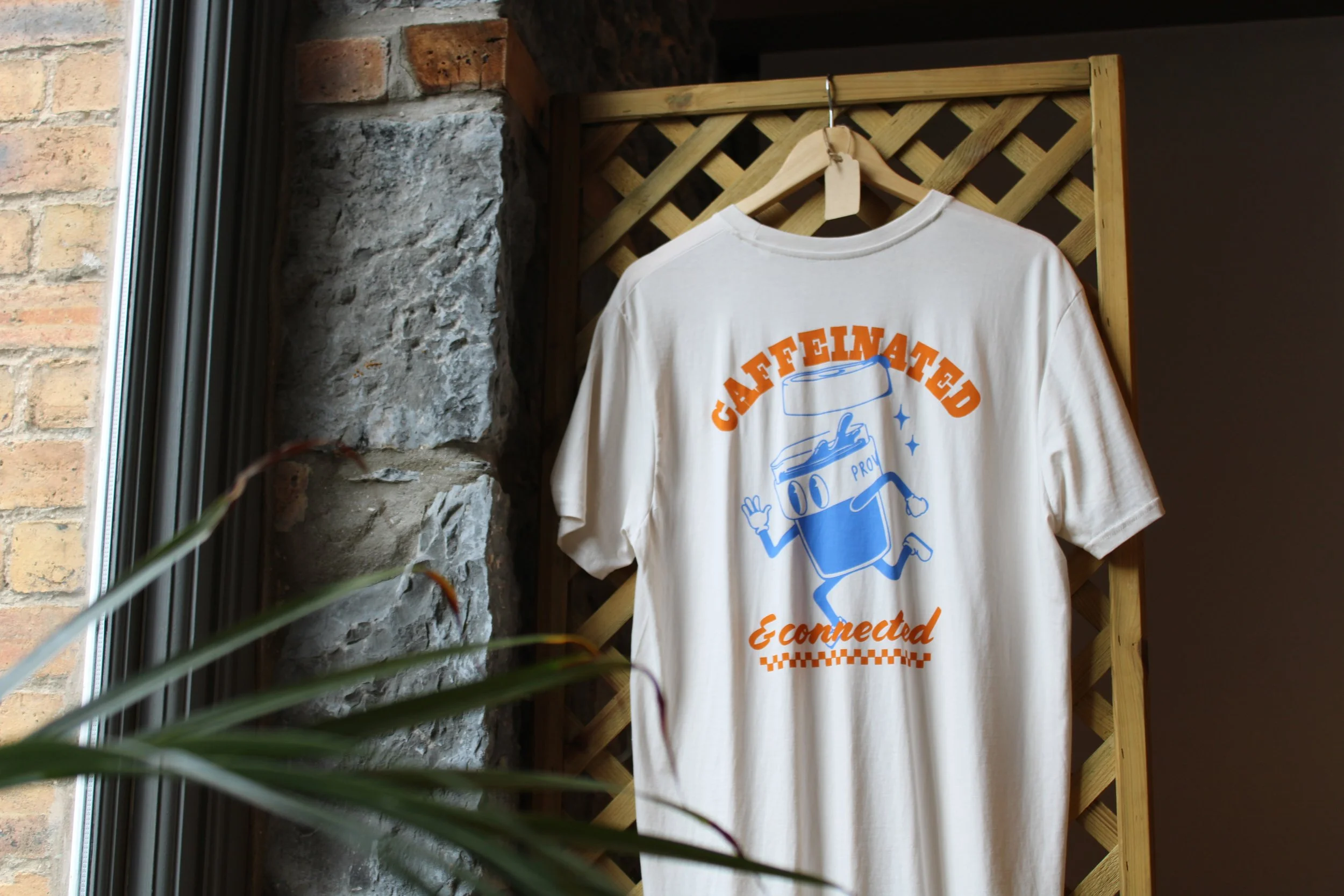A white T-shirt hangs on a wooden lattice, featuring a cartoon character of a coffee cup with arms, legs, and a smiling face. The text on the shirt reads "Caffeinated & Connected" in orange and blue colors.