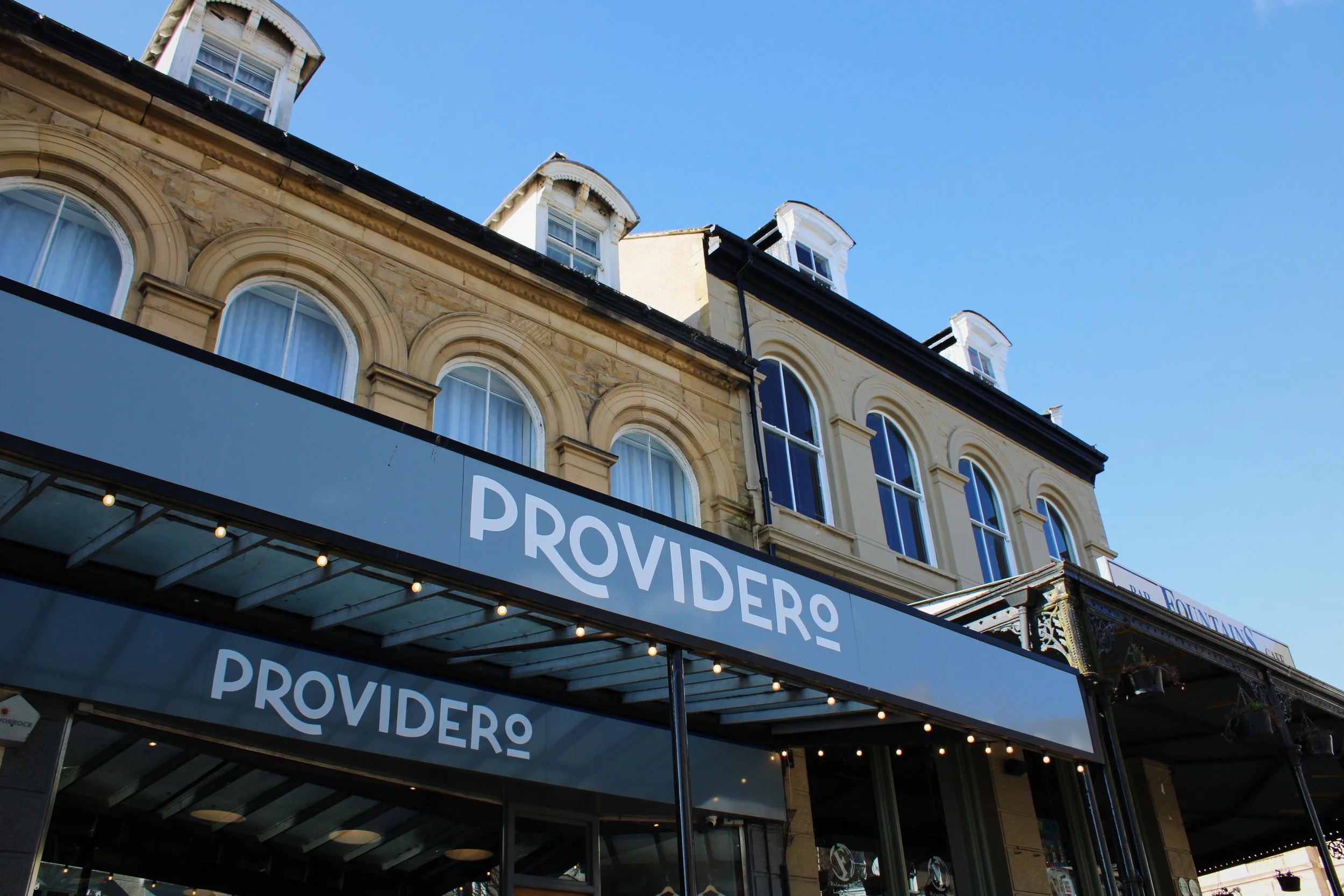 Storefront with a sign that reads 'PROVIDERO' on a black background, above a glass entrance, with classic-style buildings and windows in the background under a clear blue sky.