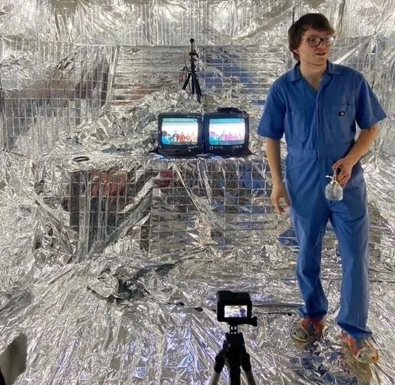 A person in blue coveralls standing in a reflective silver foil-covered room with two monitors and a camera setup.