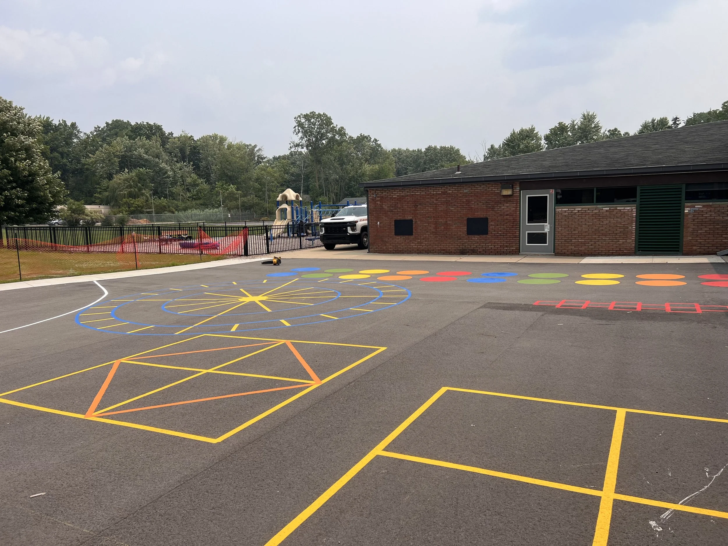 Colorful hopscotch, spider web, and circle game markings painted on asphalt outside a school building, with playground equipment and trees in the background.